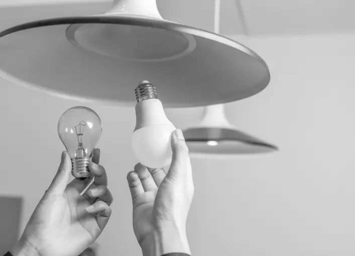 Two hands with one hand removing an old, innefficient bulb from a light fixture and a new efficient bulb about to be installed into said light fixture