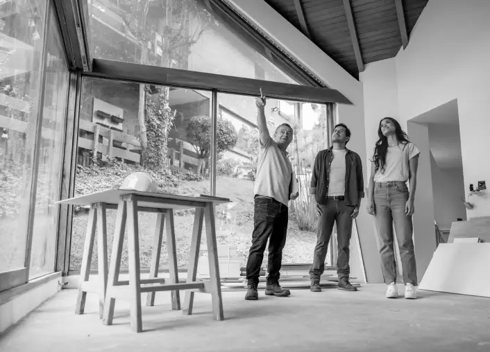 Two homeowners and a contractor within an unfinished room of a house, pointing at the ceiling