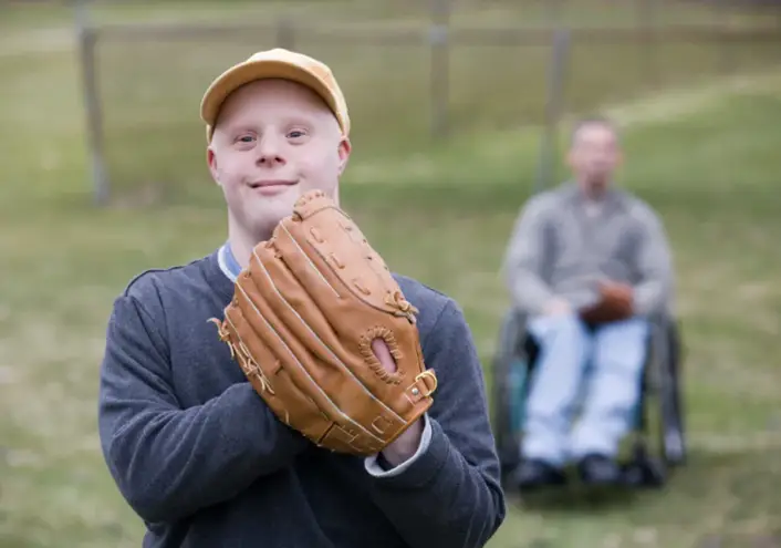 A man expericing the joy of playing baseball holding a baseball glove and with a person in a wheelchair in the background