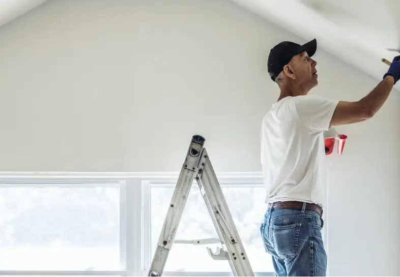 Painter on a ladder painting the wall white inside of a bright room